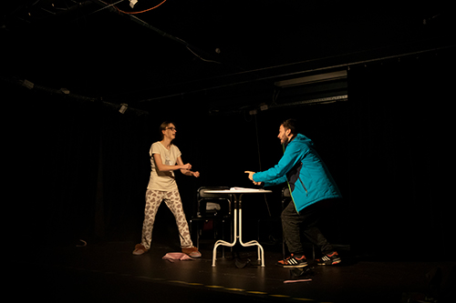 Spektakl teatralny: dwie osoby na scenie wokół okrągłego stolika.